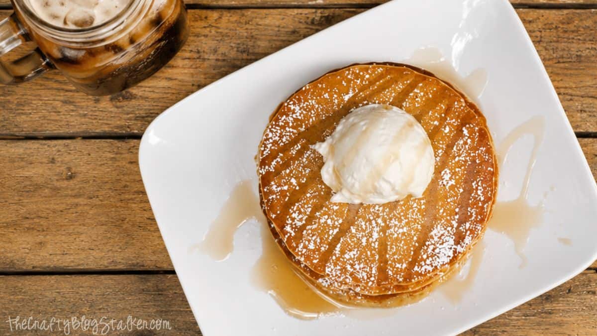 Stack of root beer pancakes topped with vanilla ice cream, powdered sugar, and syrup on a white plate beside an iced root beer drink.