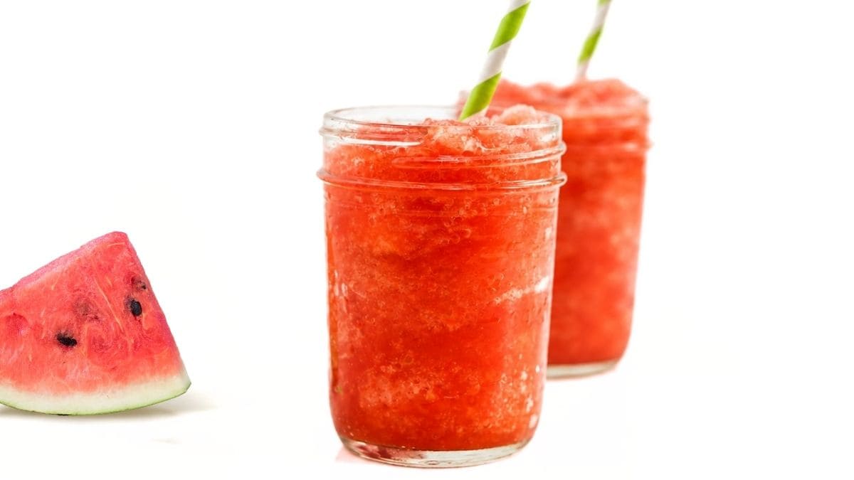 Two mason jars filled with bright red watermelon agave snowballs, served with striped paper straws and a fresh watermelon slice on a white background.