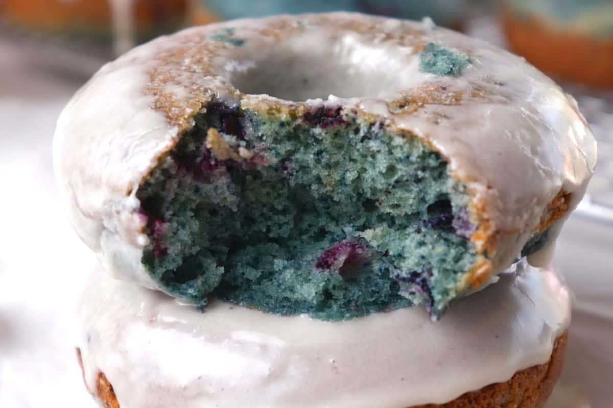 A close-up of two stacked blueberry cake donuts with white glaze, one with a bite taken out, showing a fluffy blue interior with bits of berry.