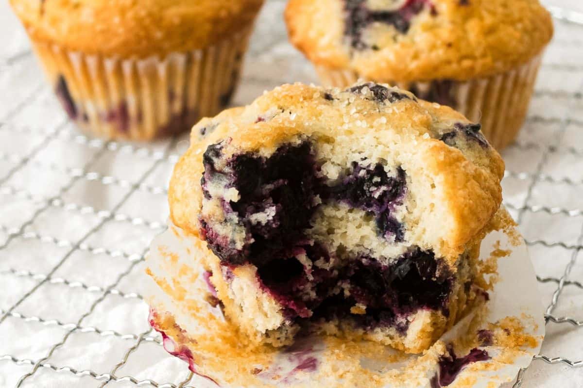 A close-up of a blueberry muffin with a bite taken out, revealing a moist, berry-filled center, set on a cooling rack with other muffins in the background.