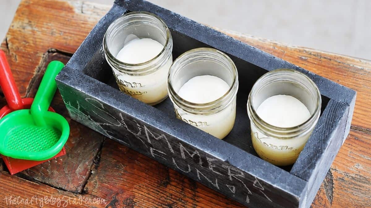 Make your very own Chalkboard Candle Box complete with DIY Citronella Candles Black wooden citronella candle box holding three mason jar candles on a rustic table beside colorful sand toys, perfect for outdoor summer entertaining.