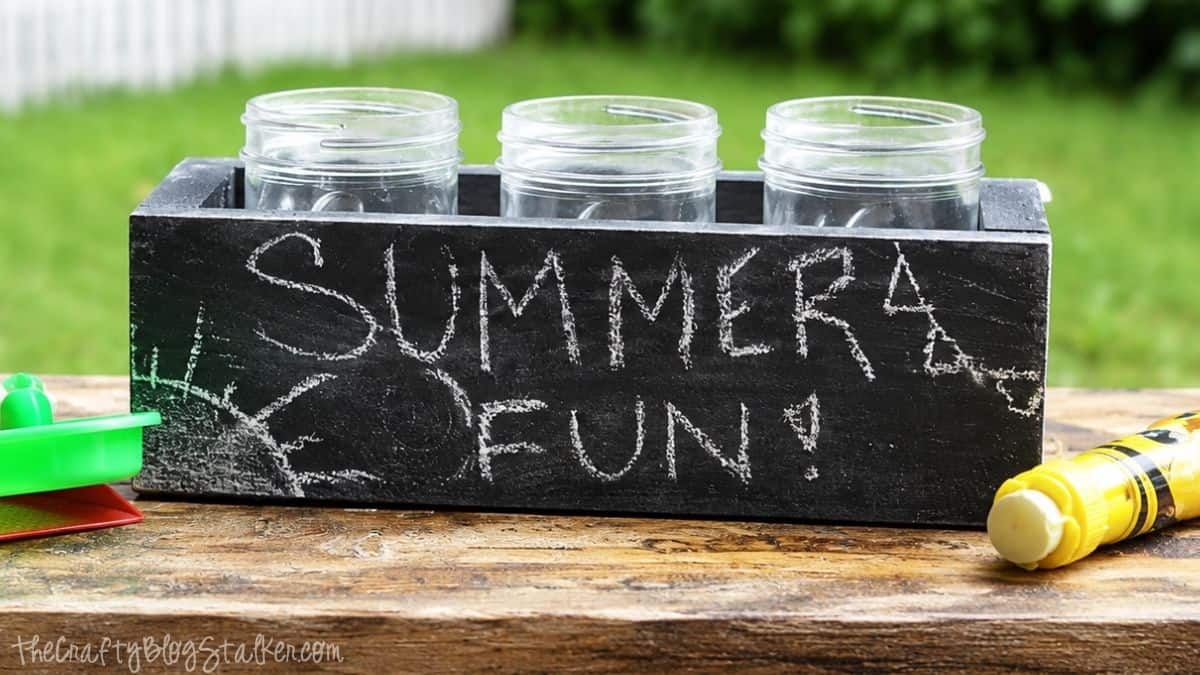 Make your very own Chalkboard Candle Box complete with DIY Citronella Candles Black wooden citronella candle box with three glass jars, chalk writing that says "Summer Fun!", and colorful sidewalk chalk on a picnic table outdoors.