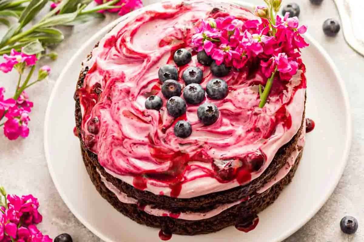 A chocolate layer cake with blueberry swirled frosting, topped with fresh blueberries and pink flowers, displayed on a white plate with scattered berries.