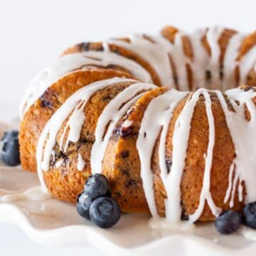 A close-up of a blueberry bundt cake drizzled with white glaze, served on a white plate and garnished with fresh blueberries.