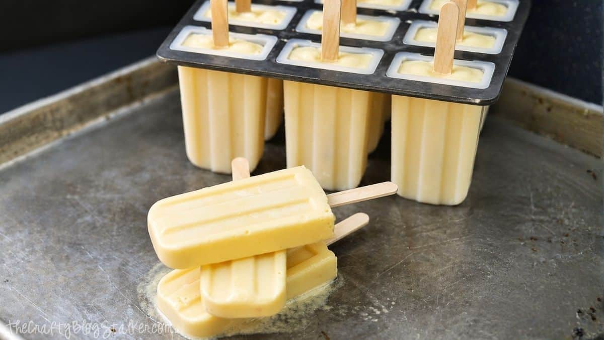 Creamy popsicles stacked on a metal tray with a filled popsicle mold behind them, each with wooden sticks and pale yellow frozen mixture.