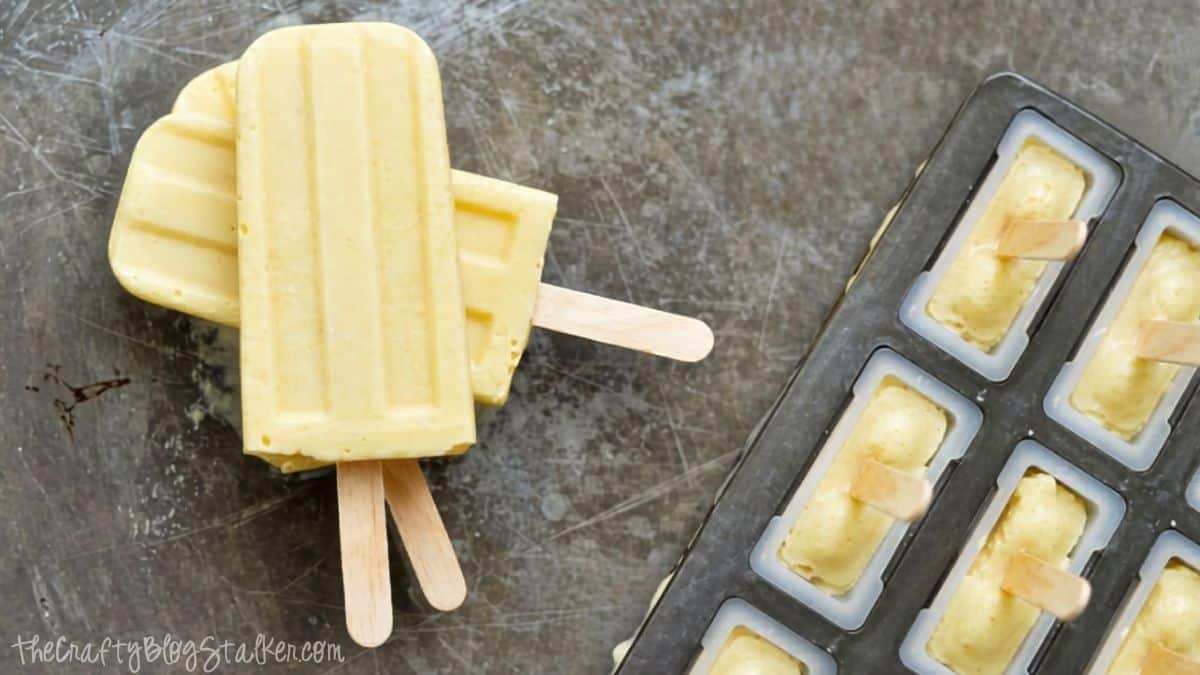 Three creamy popsicles stacked beside a popsicle mold tray filled with the same mixture on a metal surface.