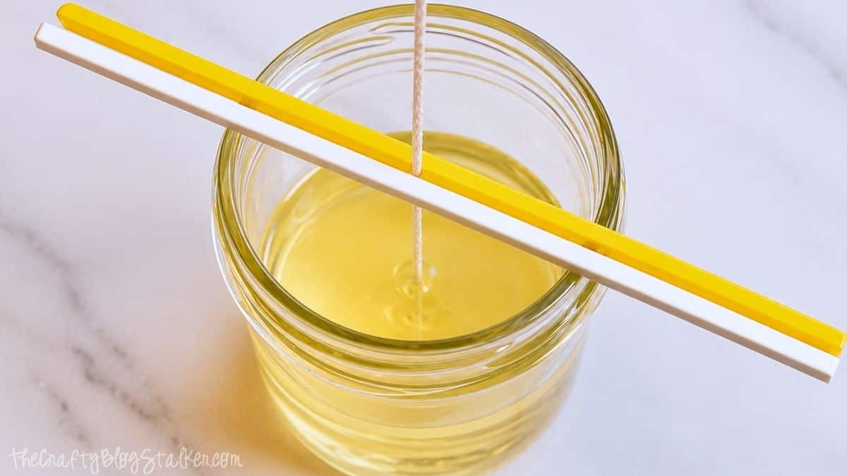 Glazen pot gevuld met gesmolten bijenwas en citronellaolie, met de pit in het midden en aan de bovenkant vastgezet met hobbystokjes.