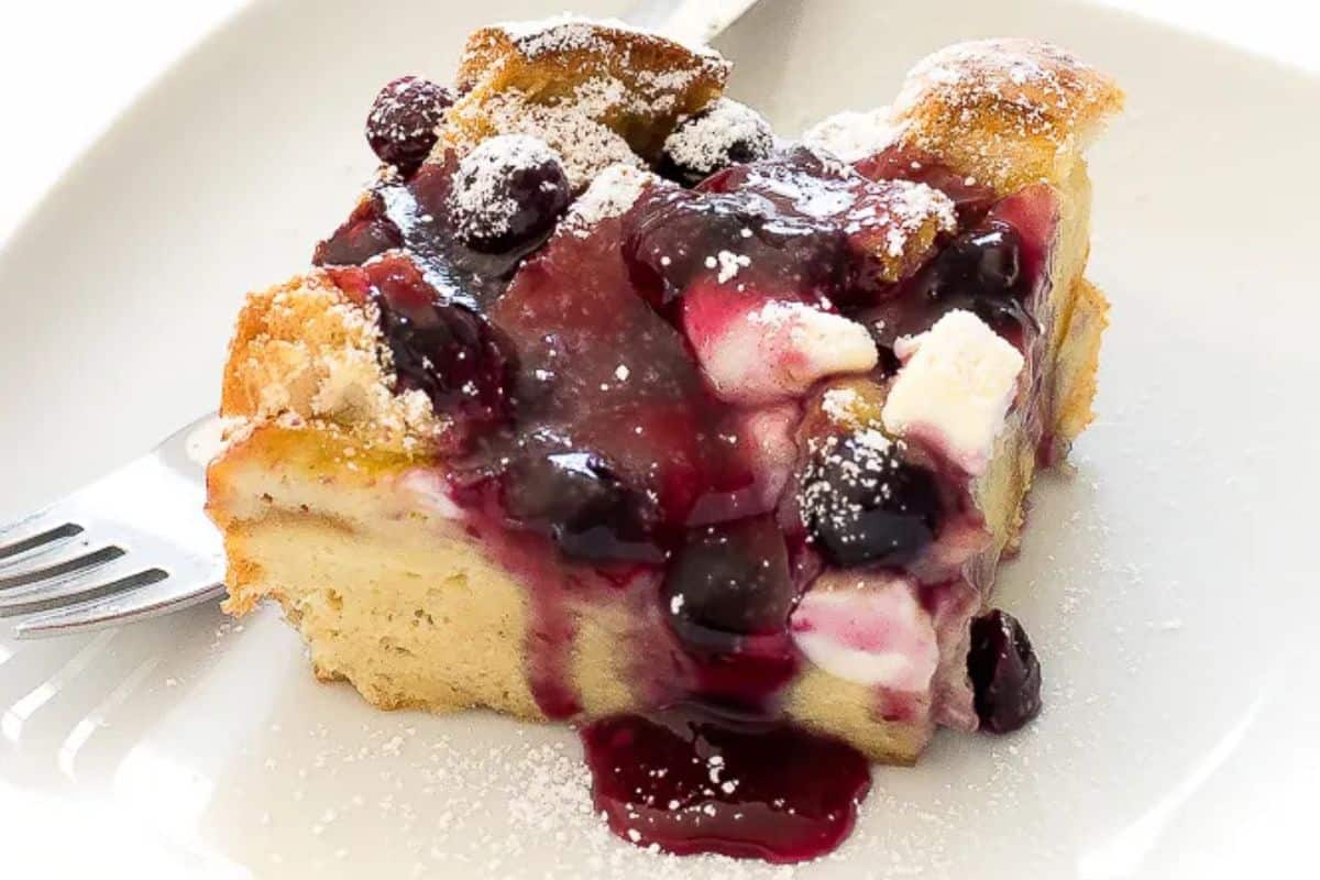 A slice of baked overnight blueberry French toast topped with blueberry sauce, cream cheese chunks, and a dusting of powdered sugar on a white plate.