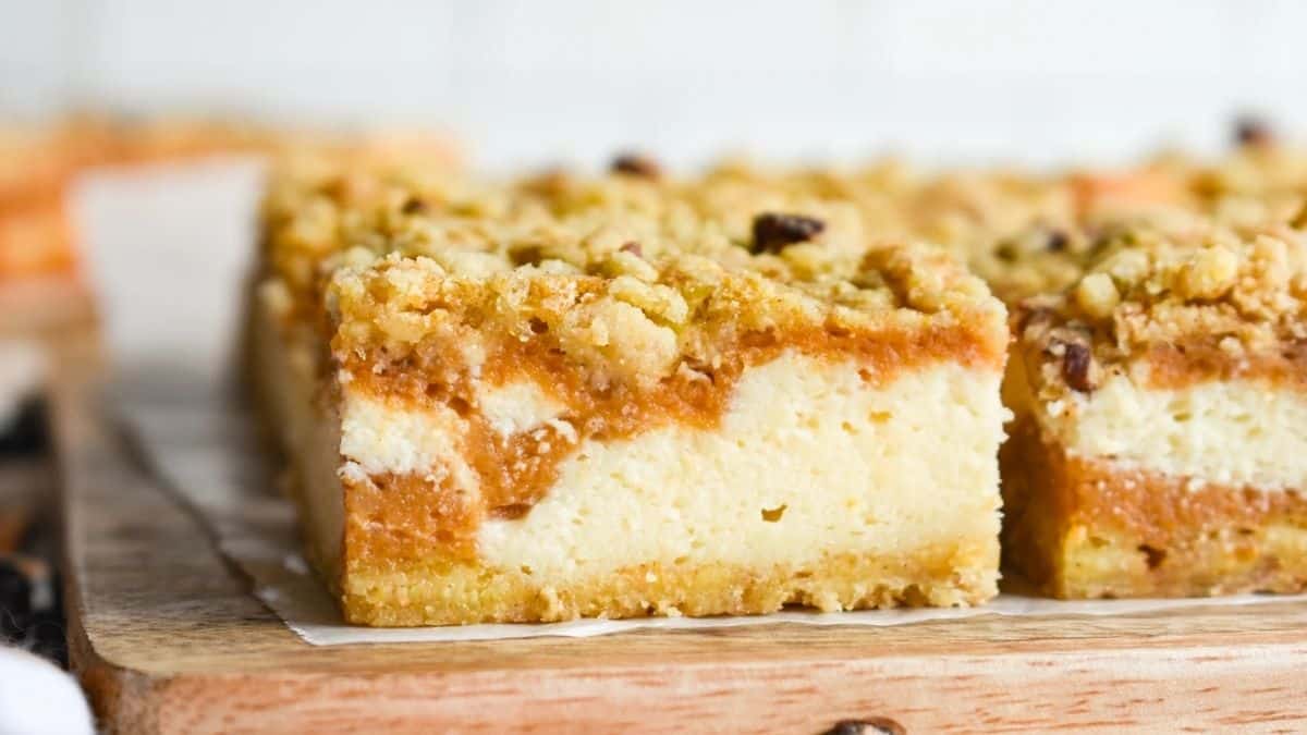 Close-up of pumpkin cheesecake bars with a crumbly streusel topping, creamy cheesecake layer, and golden pumpkin filling on a wooden board.