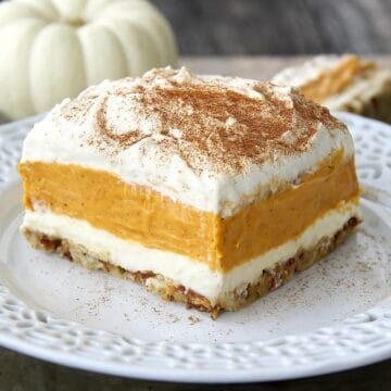 Slice of layered pumpkin dessert with creamy pumpkin filling, whipped topping, and a nutty crust, sprinkled with cinnamon on a white plate.