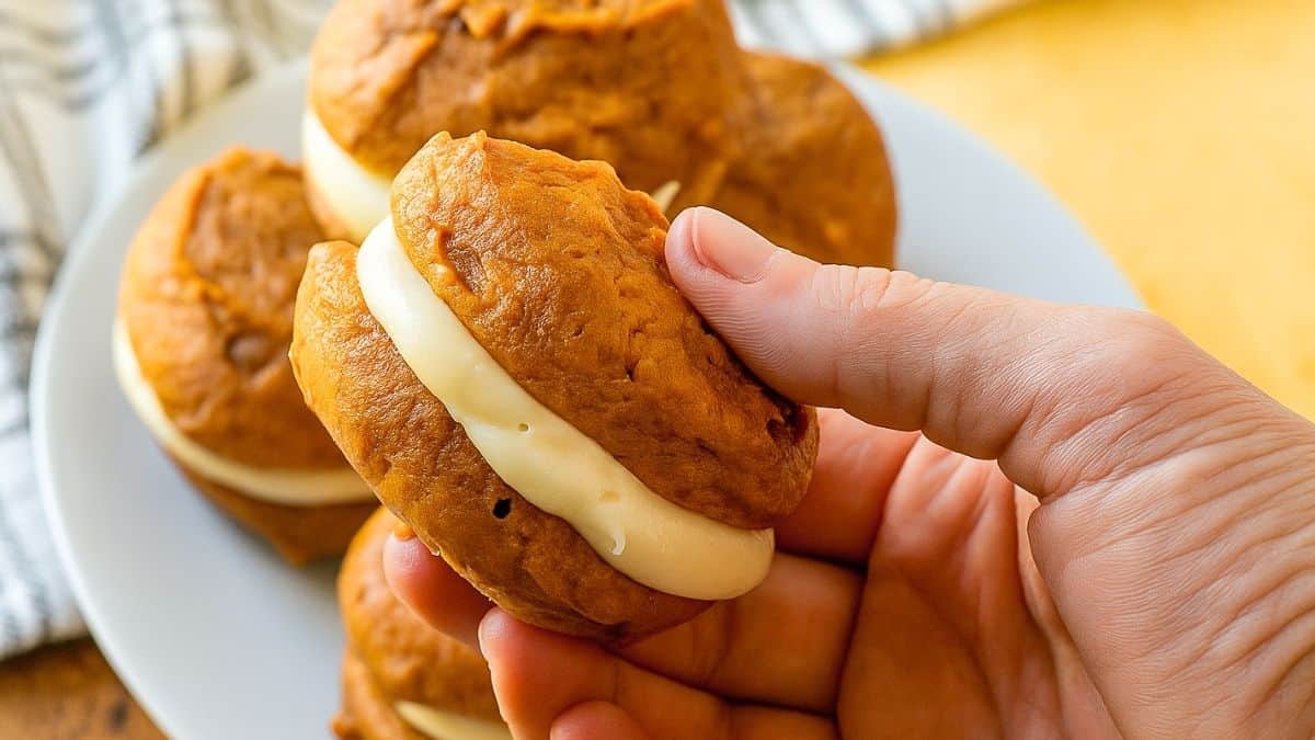 Hand holding a pumpkin whoopie pie filled with creamy frosting, with more pumpkin sandwich cookies on a white plate in the background.