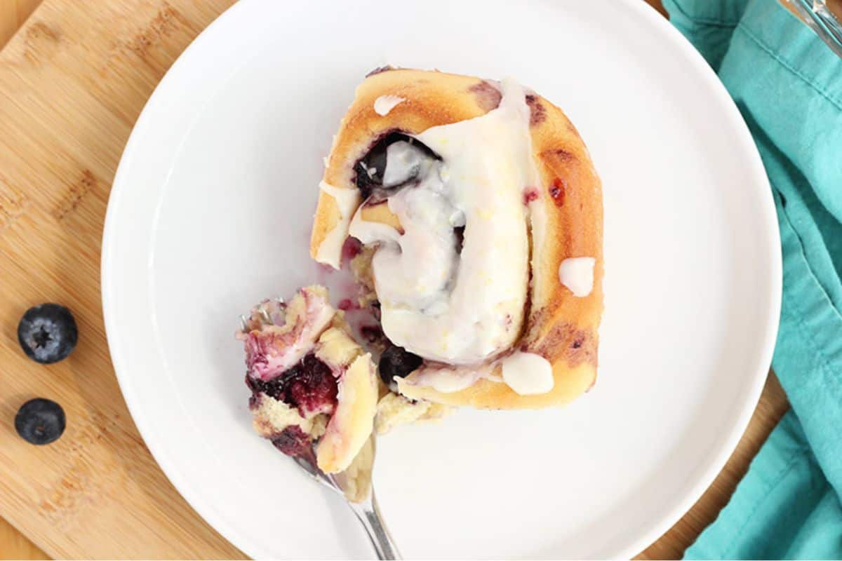 A blueberry lemon sweet roll topped with creamy icing, served on a white plate with a bite on a fork and fresh blueberries on the side.
