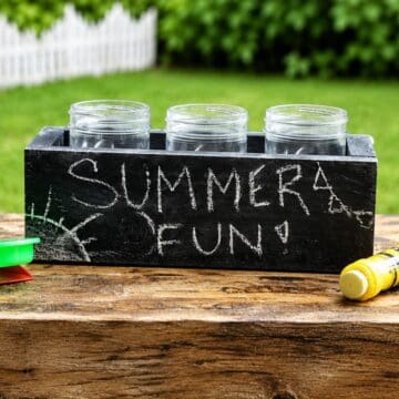Black wooden box holding three mason jars with "Summer Fun!" written in chalk, displayed on a rustic outdoor table with a green lawn in the background.