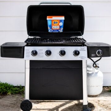 Gas grill with open lid and a container of Tide Oxi cleaner placed on the grill grate outside on a patio.