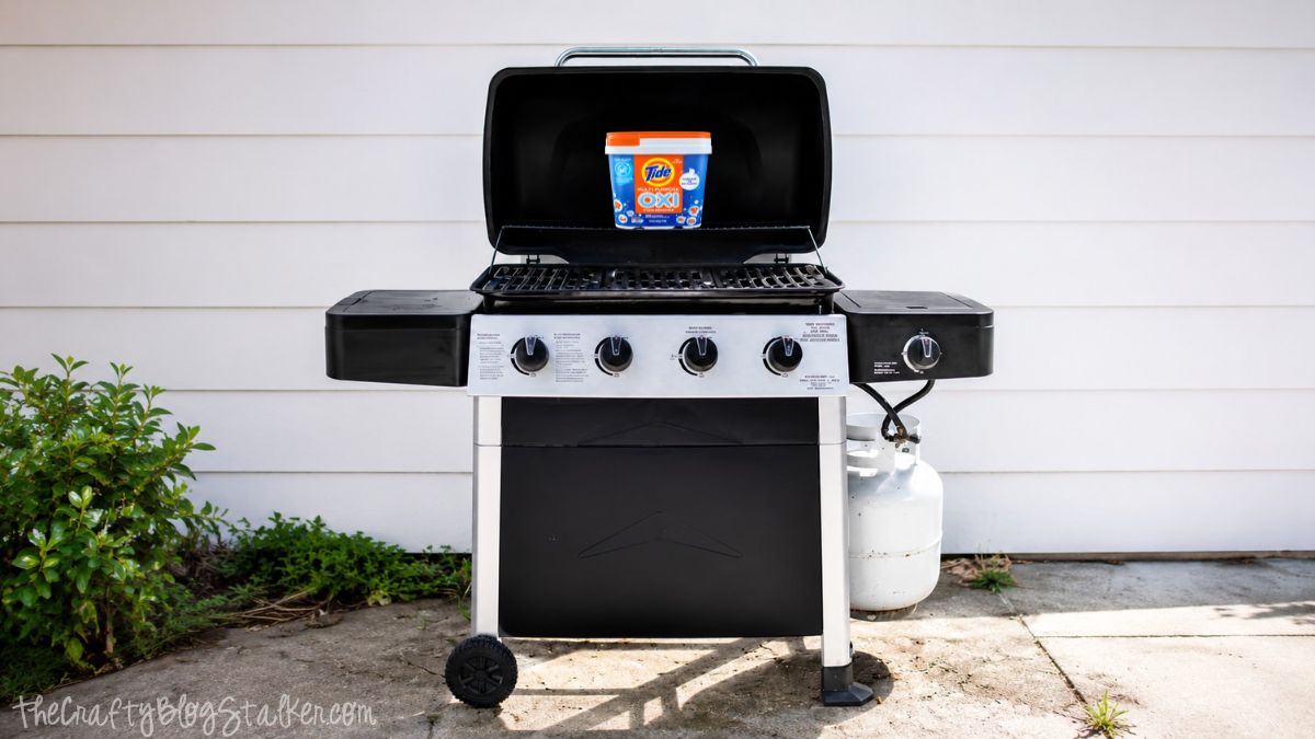 Do you have a gross grill? Learn How to Clean your BBQ Grill fast and easy! Gas grill with open lid and a container of Tide Oxi cleaner placed on the grill grate outside on a patio.