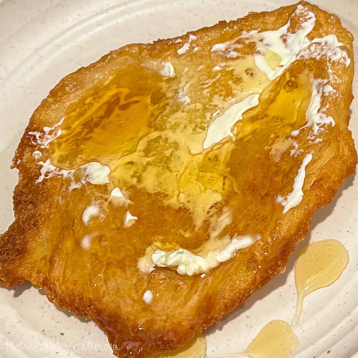 Close-up view of a golden-brown piece of fry bread topped with melted butter and drizzled honey, sitting on a speckled beige plate. The fry bread has a crispy, bubbly texture with glistening spots of honey and soft butter, creating an irresistible, indulgent look. Perfect comfort food for a delicious treat!