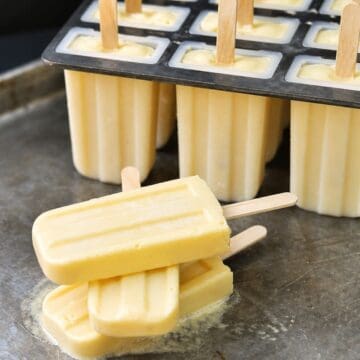 Creamy popsicles stacked on a metal tray with a filled popsicle mold behind them, each with wooden sticks and pale yellow frozen mixture.