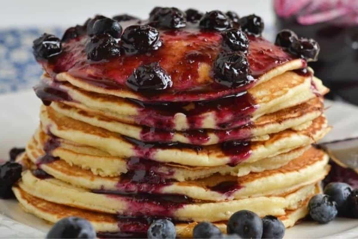 Stack of fluffy pancakes topped with warm homemade blueberry syrup and whole blueberries, served on a white plate with extra berries on the side.