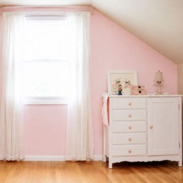 Pink painted accent wall with a tall window, white lace curtains, hardwood floors, and a white dresser decorated with vintage accessories.