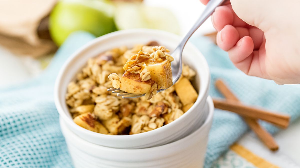 Hand holding a forkful of warm apple crumble with oats and cinnamon, served in a white ramekin on a blue cloth.