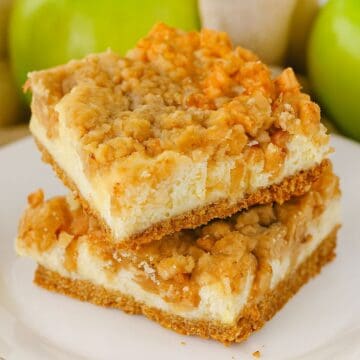 Two apple cheesecake bars stacked on a white plate, featuring a crumbly streusel topping and graham cracker crust with green apples in the background.