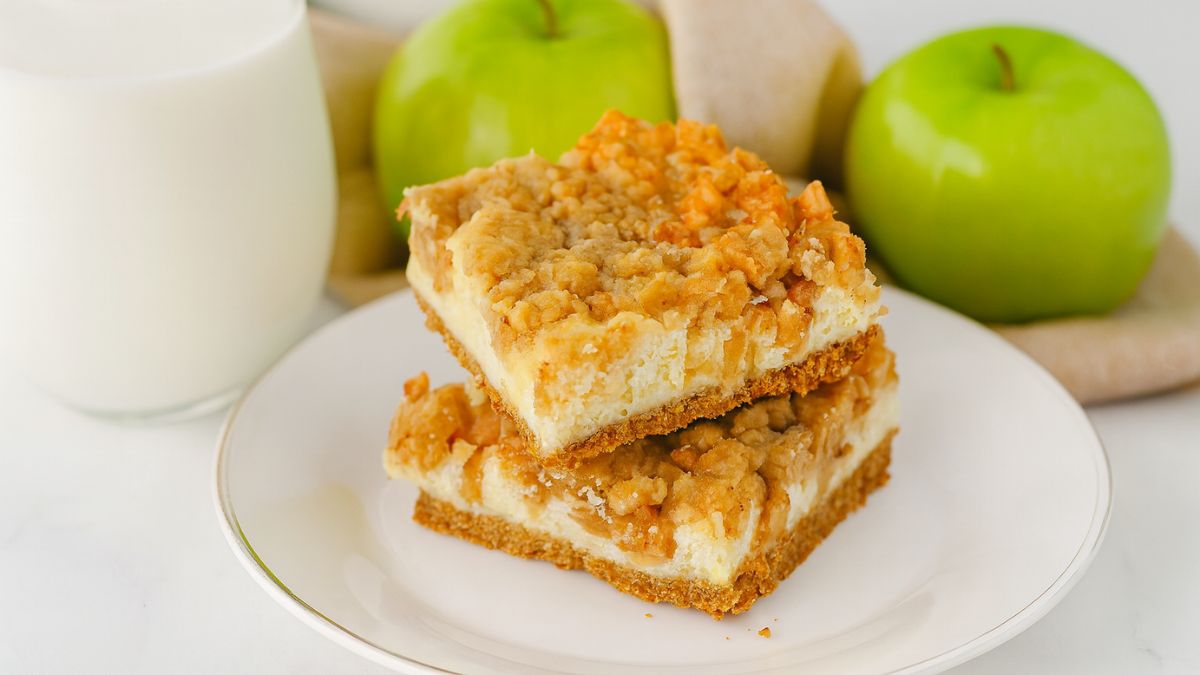 Two apple cheesecake bars stacked on a white plate, topped with crumbly streusel and served beside a glass of milk and green apples.