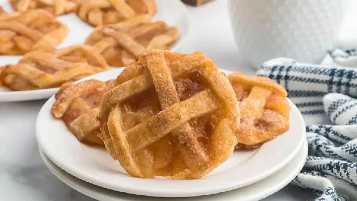 Small lattice-topped apple pie cookies stacked on white plates with more cookies and a blue-striped napkin in the background.
