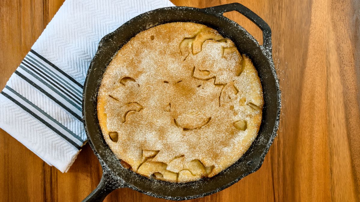 Golden apple puff pancake baked in a cast iron skillet, dusted with powdered sugar, on a wooden table beside a striped kitchen towel.