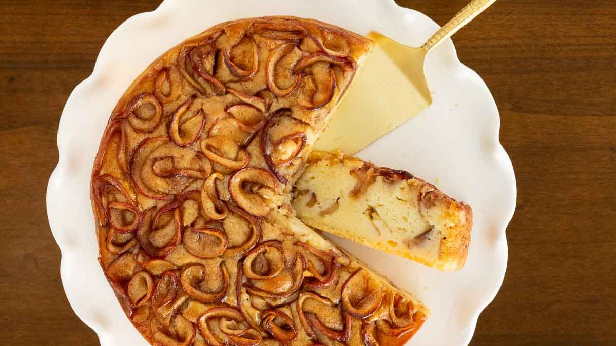 Golden apple ricotta cake on a white platter with one slice removed, showing moist layers of baked apples and creamy ricotta inside.