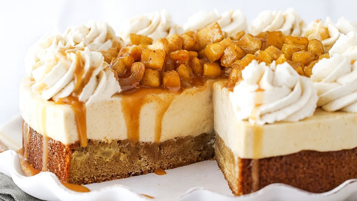 Caramel apple blondie cheesecake topped with spiced apple chunks, caramel drizzle, and whipped cream, with a slice removed to show layers.