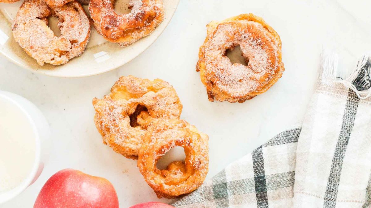 Golden apple fritter rings coated in cinnamon sugar on a white surface with fresh apples and a plaid napkin beside them.