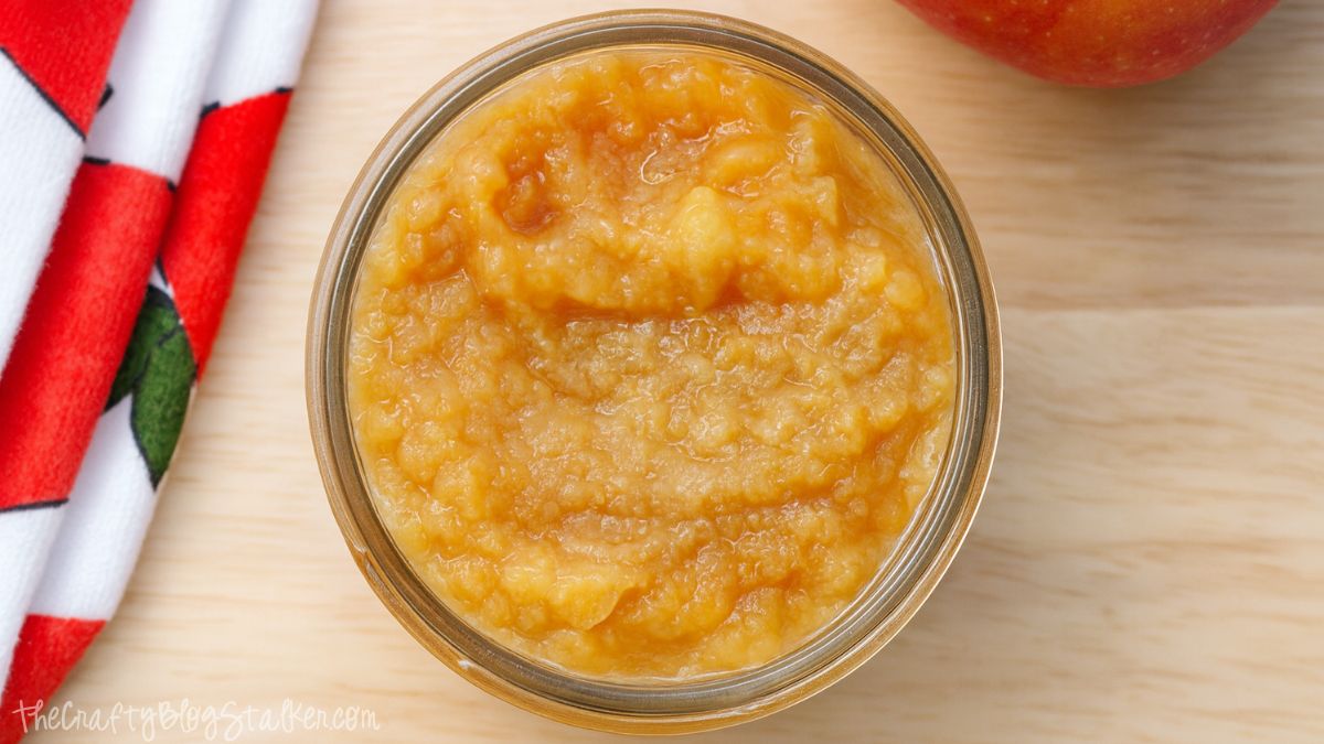 Jar of homemade applesauce with a smooth, golden texture on a wooden surface beside a red apple and a festive towel.
