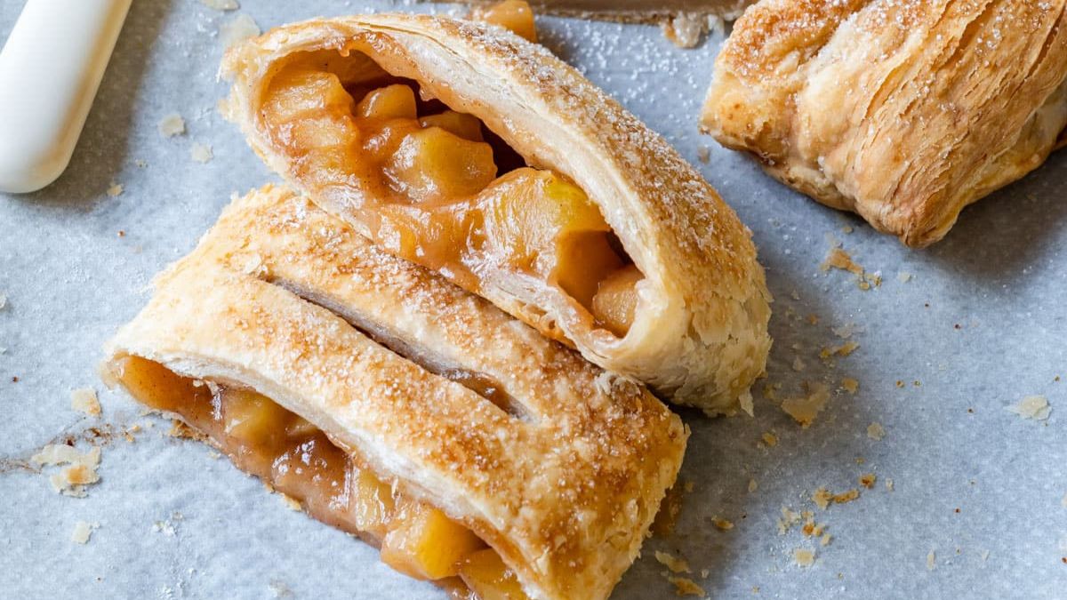 Flaky puff pastry apple strudel sliced open, showing warm cinnamon-spiced apple filling on a baking sheet.