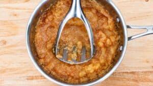 Homemade unsweetened applesauce being mashed with a potato masher in a stainless steel saucepan on a wooden cutting board.