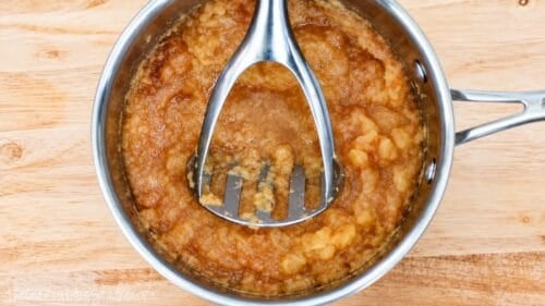Homemade unsweetened applesauce being mashed with a potato masher in a stainless steel saucepan on a wooden cutting board.