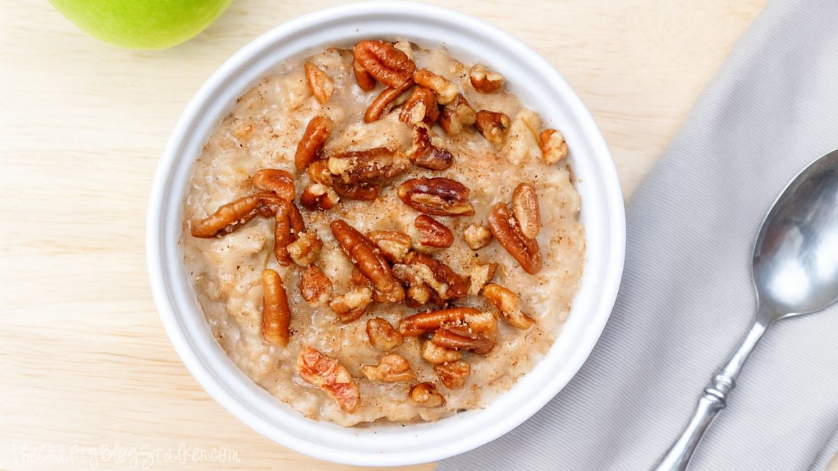 Bowl of warm apple pie oatmeal topped with chopped pecans, set on a wooden table beside a spoon and a green apple.
