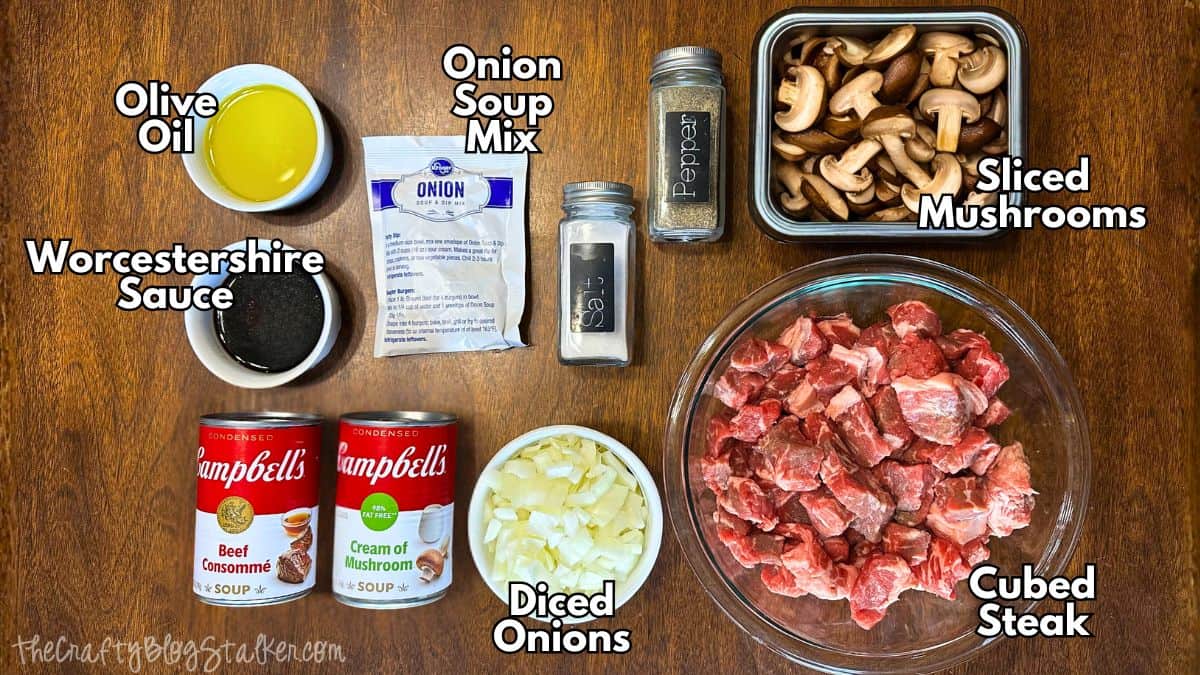 Labeled ingredients for slow cooker beef tips including cubed steak, sliced mushrooms, diced onions, onion soup mix, beef consommé, cream of mushroom soup, olive oil, Worcestershire sauce, salt, and pepper laid out on a table.
