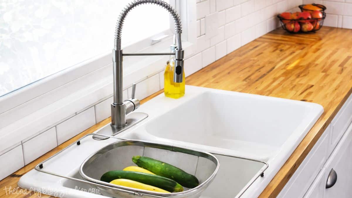 Farmhouse sink with a tall spring faucet set in butcher block countertops, with a metal colander holding cucumbers and squash on the sink.