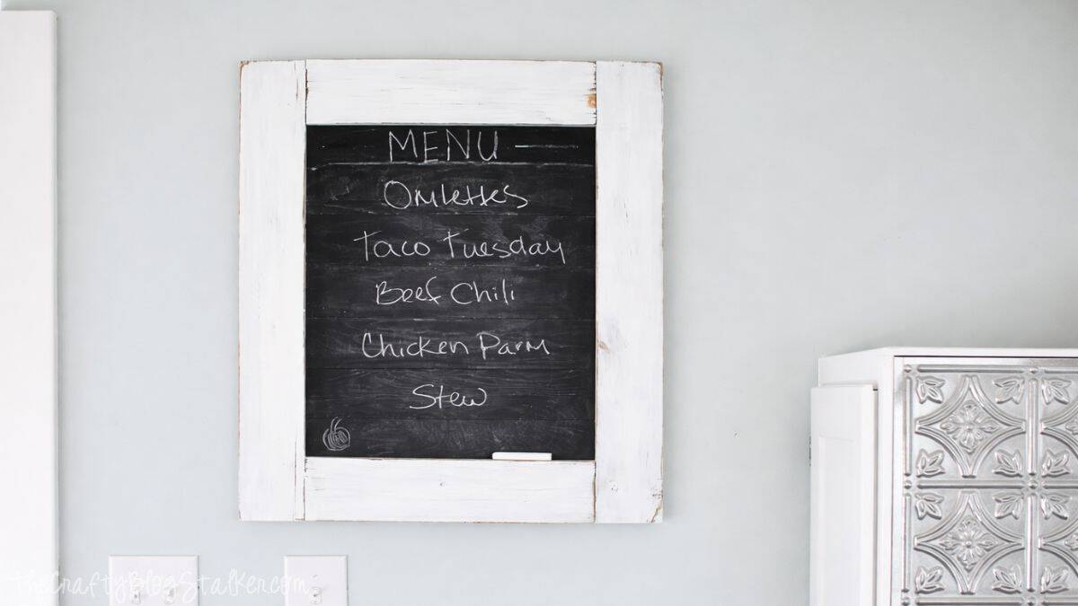 Farmhouse chalkboard menu with a white wood frame listing meals like omelettes, taco Tuesday, beef chili, chicken parm, and stew.
