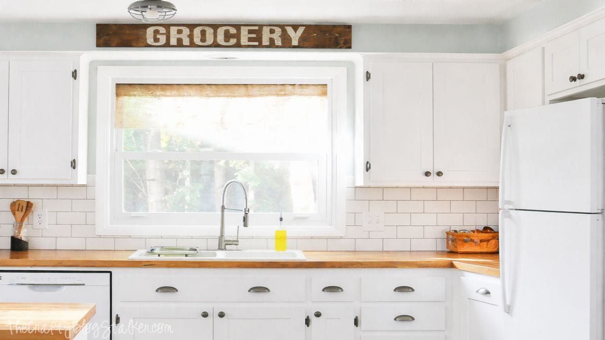 Bright farmhouse kitchen with white cabinets, butcher block countertops, subway tile backsplash, farmhouse sink under a window, and a wooden Grocery sign above.