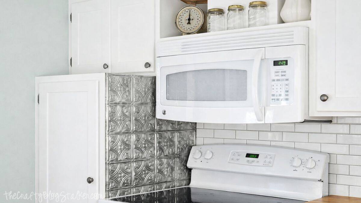 White microwave above a stove in a farmhouse kitchen with white cabinets, subway tile backsplash, and decorative metal backsplash tiles behind the range.