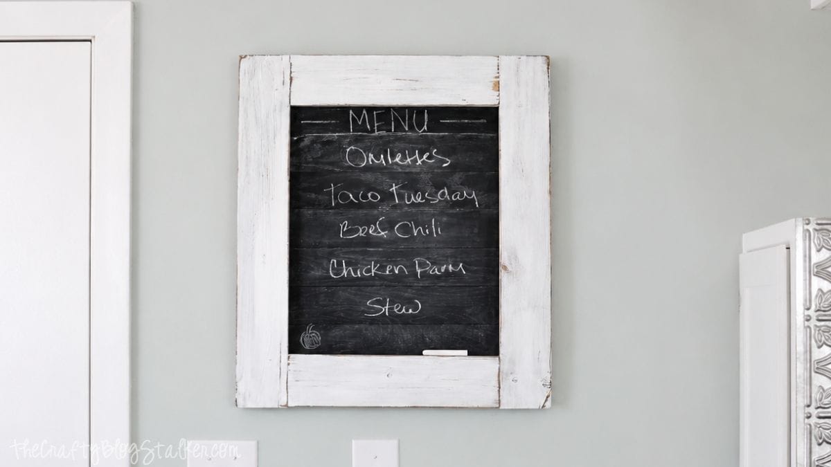 Is the family always asking what's for dinner? Create this super cute planked Kitchen Chalkboard Menu to keep them up to date! Click through for the tutorial on how to make this chalkboard menu! Rustic framed chalkboard menu hanging on a gray kitchen wall with a distressed white wood frame and handwritten weekly meal ideas.