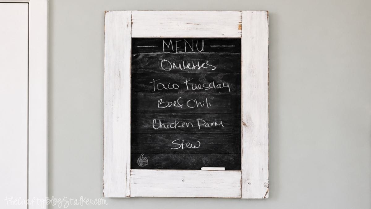 Is the family always asking what's for dinner? Create this super cute planked Kitchen Chalkboard Menu to keep them up to date! Click through for the tutorial on how to make this chalkboard menu! Rustic framed chalkboard menu board hanging on a gray wall with a distressed white wood frame and handwritten weekly dinner ideas.