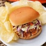Slow cooker BBQ shredded chicken sandwich topped with creamy coleslaw, served on a bun with potato chips on a white plate.