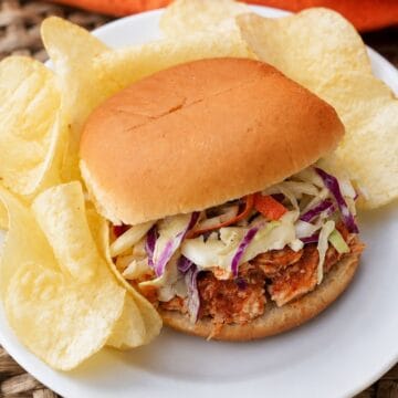 Slow cooker BBQ shredded chicken sandwich topped with creamy coleslaw, served on a bun with potato chips on a white plate.