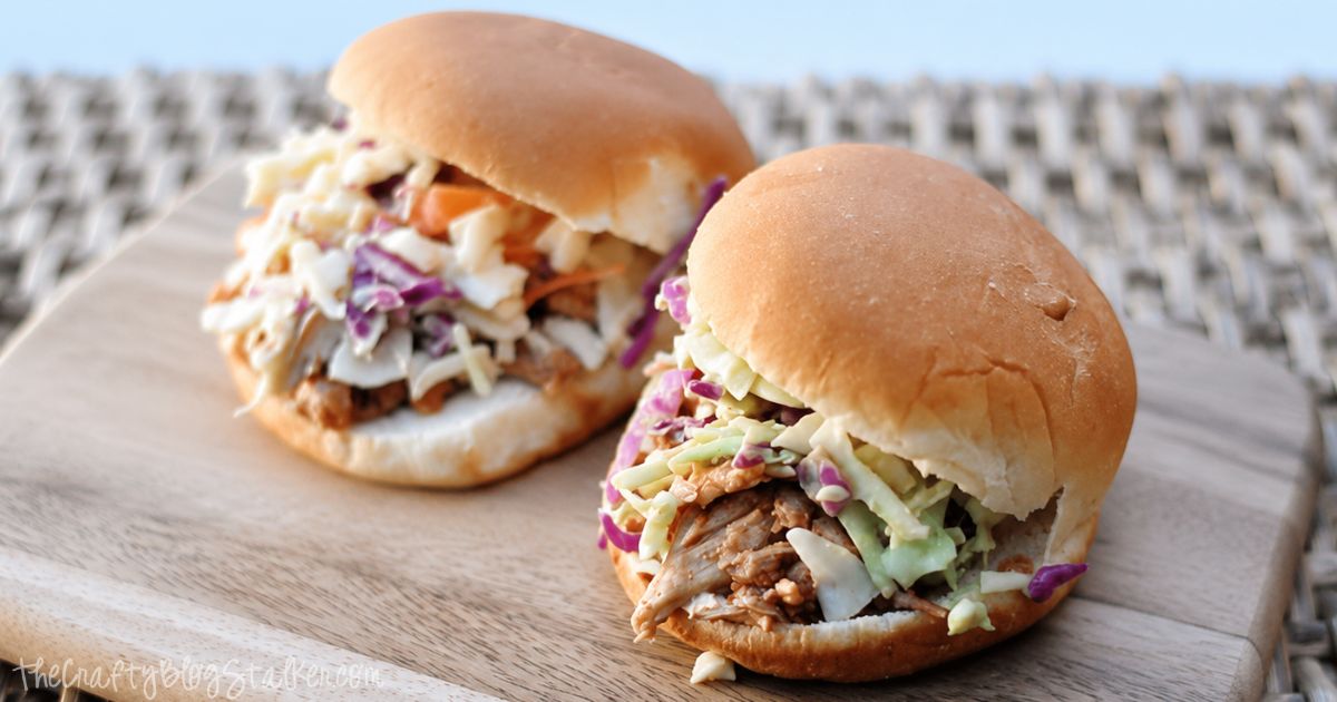Two BBQ shredded chicken slider sandwiches topped with creamy coleslaw on soft buns, served on a wooden board.