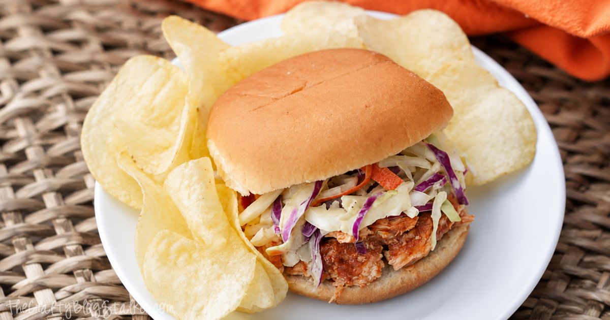 BBQ shredded chicken sandwich topped with coleslaw on a bun, served with potato chips on a white plate.