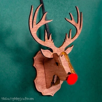 Wooden 3D reindeer head ornament with antlers and a red pom-pom nose hanging against a green background.