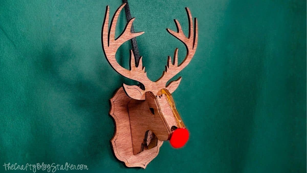 3D wooden reindeer ornament with large antlers and a bright red pom-pom nose hanging against a dark green background.
