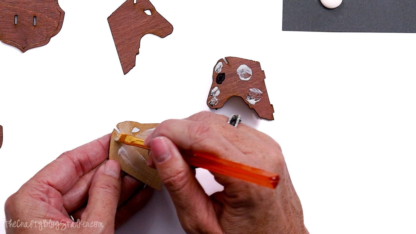 Hands applying wood glue with a small brush to a laser-cut wooden reindeer piece, with other ornament parts and glue drops visible on the workspace.